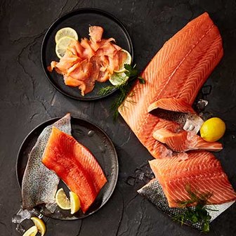 Various cuts of raw salmon with lemon slices on black plates on a slate tabletop.