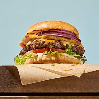 Close-up of a burger with lettuce, tomato, beef patty, cheese, red onion, and sauce in a toasted bun, placed on parchment over a wooden surface.