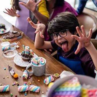  A wooden table is covered with vibrant treats. lollipop sits on a white plate. Scattered around are small round candies, a spoon,    and more marshmallows. In the background, additional desserts like a chocolate cake    are visible. Several people are se
