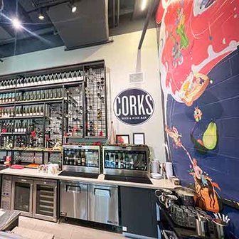 Interior of Corks Beer & Wine Bar with shelves of wine bottles, bar equipment, and a colorful mural on the wall.