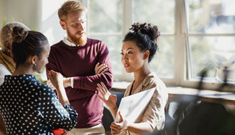 Three people standing and talking in a bright indoor space near large windows, with one person holding a tablet or document