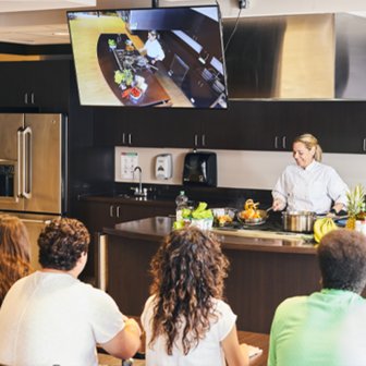 Chef giving a cooking demonstration in a modern kitchen while attendees watch, with overhead screen showing the prep area.