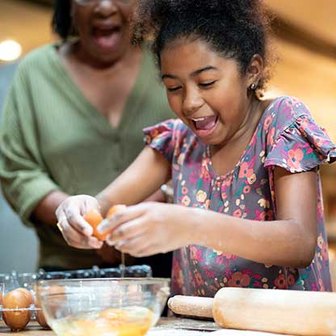 Child cracking an egg into a glass bowl on a floured surface with rolling pins nearby.