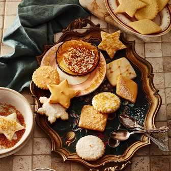 Assorted holiday cookies in star and square shapes decorated with white and gold sprinkles, arranged on an ornate tray with a bowl of sprinkles and silver spoons.