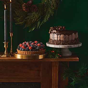Holiday dessert display with a chocolate cake topped with nuts on a marble stand and a berry tart on a gold tray, set beside black candles and festive greenery.