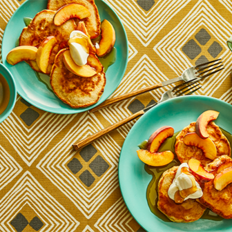 Plates of pancakes topped with peach slices, whipped cream, and syrup on a patterned tablecloth.