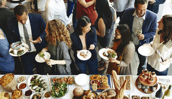 People standing and holding plates of food around a long table filled with assorted breads, fruits, and appetizers.