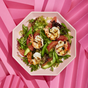 Salad with grilled shrimp, grapefruit segments, avocado slices, and mixed greens served on a pink plate against a pink folded paper background.
