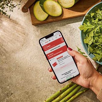 Hand holding a smartphone showing Thank You Rewards offers, with sliced zucchini, leafy greens, and asparagus on a cutting board in the background.