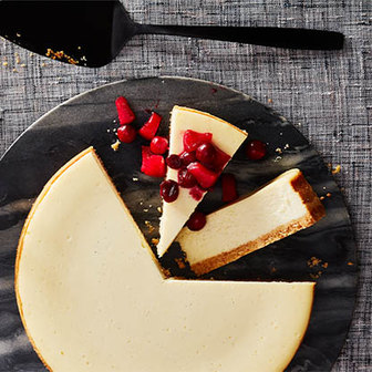 Cheesecake with a slice topped with apple and cranberry pieces on a dark serving board with a cake server.