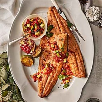 Cooked salmon fillets on a platter topped with diced vegetables and herbs, served with roasted lime and pomegranate.