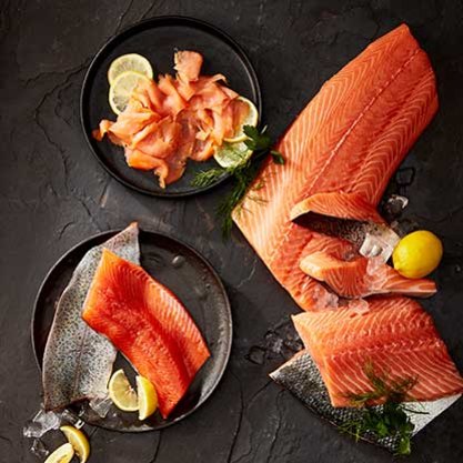 Various cuts of raw salmon with lemon slices on black plates on a slate tabletop.