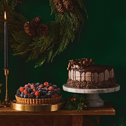 Holiday dessert table featuring a chocolate layer cake and mixed berry tart beneath festive evergreen décor.