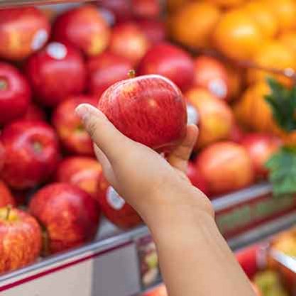 Hand holding a red apple in front of a display of fresh apples at a grocery store.