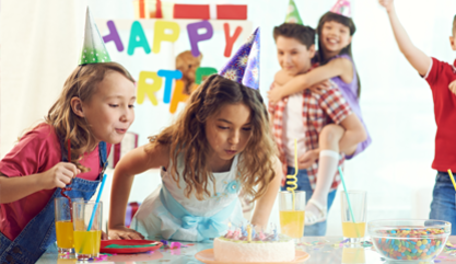 Children wearing party hats gathered around a table with a birthday cake, drinks, and colorful decorations.