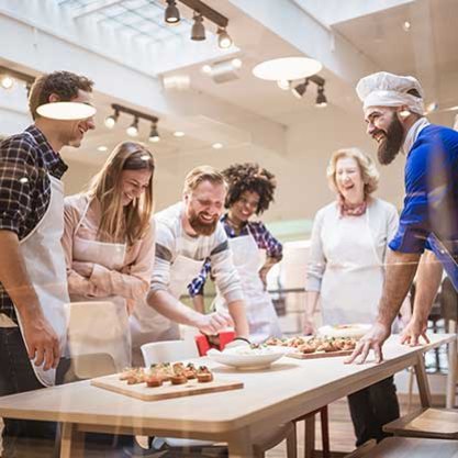 Group of people wearing aprons gathered around a table with prepared food during a cooking class.