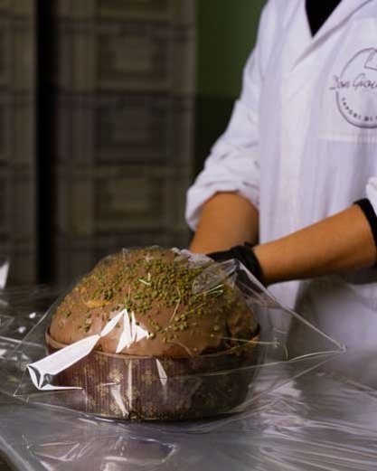 Person in white coat wrapping a chocolate-glazed panettone in clear plastic.