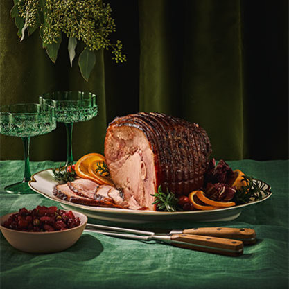 Glazed holiday ham on a platter with orange slices and herbs, served with cranberry sauce and two green glasses on a green tablecloth.