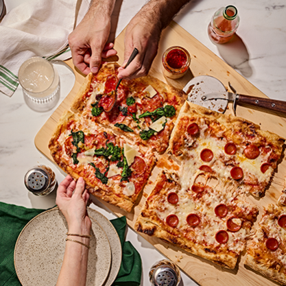Two rectangular pizzas on a wooden board, one topped with pepperoni and the other with spinach, tomato, and cheese, with hands reaching for slices and drinks nearby.