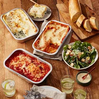 Three trays of baked lasagna with a side salad, sliced bread, and glasses of drinks on a wooden table.