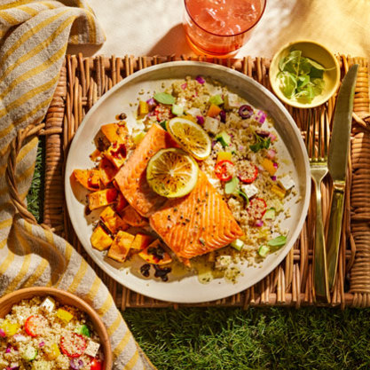 Plate of grilled salmon with lemon slices served over quinoa salad with vegetables, alongside a bowl of extra salad, a drink, and a small dish of herbs on a picnic-style setup.