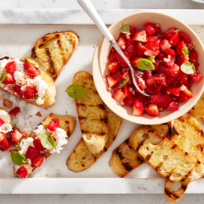 Grilled bread slices topped with ricotta, strawberries, and basil next to a bowl of strawberry tomato mixture.