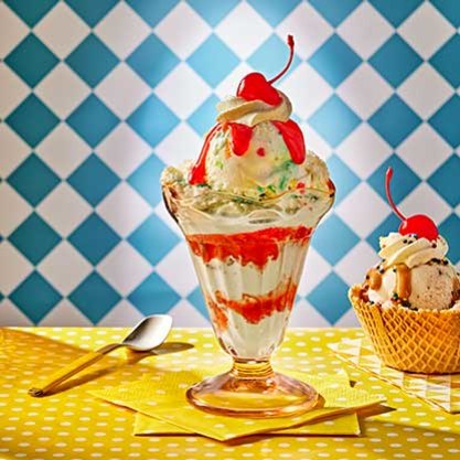 Ice cream sundae in a glass dish topped with whipped cream, cherry, and strawberry sauce, with a waffle bowl sundae in the background on a yellow polka-dot table.