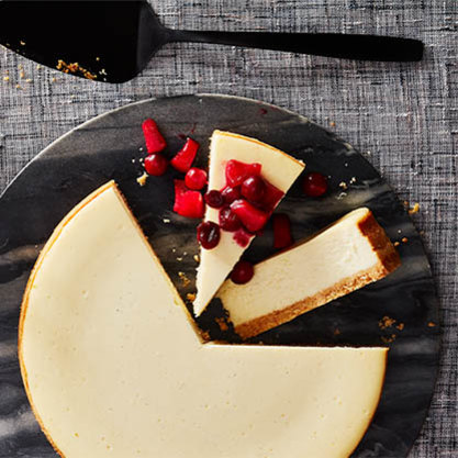 Cheesecake with a slice topped with apple and cranberry pieces on a dark serving board with a cake server.