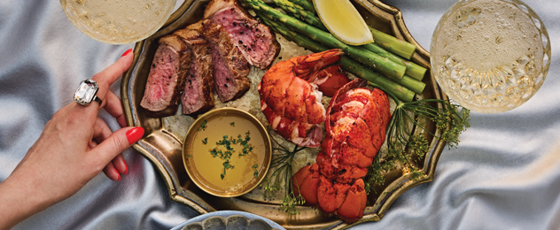 Elegant surf and turf platter with sliced steak, lobster tail, asparagus, lemon wedge, and butter sauce.
