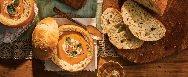 Rustic artisan bread loaves and sliced sourdough surrounding a bowl of creamy soup served in a bread bowl.