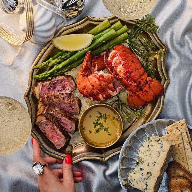 Elegant surf and turf platter with lobster tail, sliced steak, asparagus, lemon wedge, and dipping sauce, served with cheese and crackers on a silver tray.