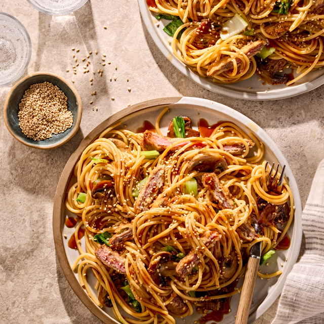 Plate of spaghetti with sliced beef, bok choy, soy sauce drizzle, and sesame seeds on a light stone table.