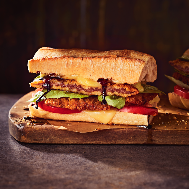 Close-up of a crispy veal Caprese sandwich with melted cheese, fresh tomato, lettuce, and balsamic glaze on rustic bread.