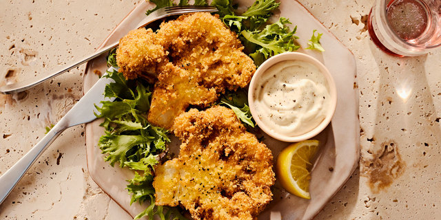 Plate of crispy breaded cauliflower steaks on greens with creamy dipping sauce and lemon wedge.