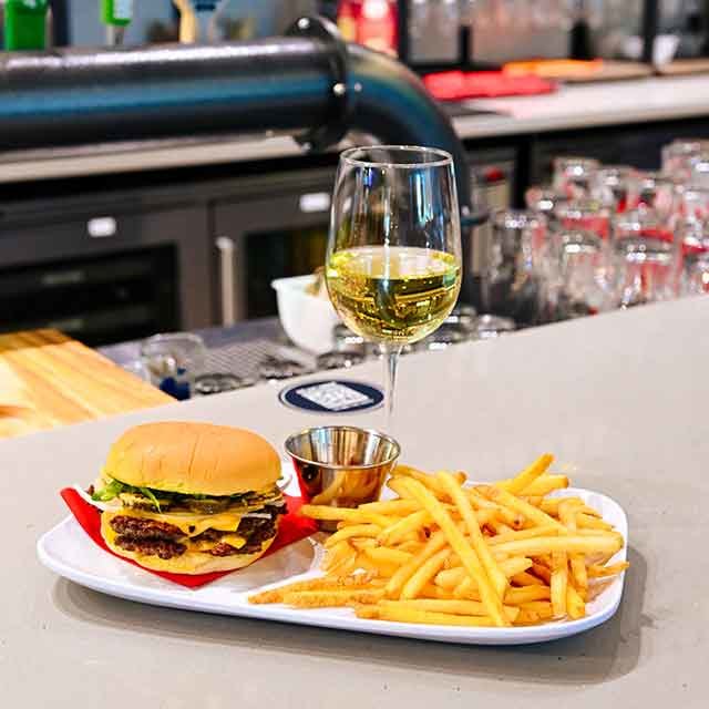 Plate with a cheeseburger, French fries, and dipping sauce next to a glass of white wine on a bar counter.