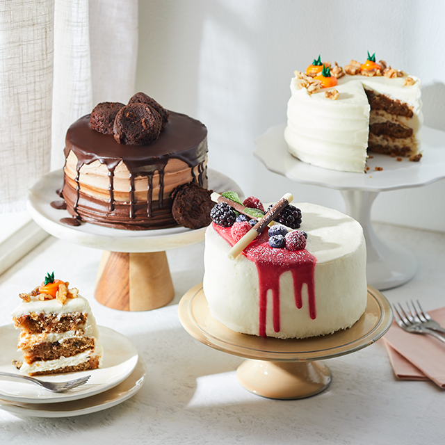 Three decorated cakes on stands and a slice on a plate, including a chocolate cake with brownie pieces, a white cake with red berry drizzle and fresh berries, and a carrot cake with nuts and frosting.