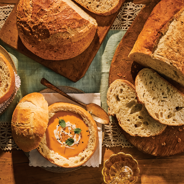 Rustic artisan bread loaves and sliced sourdough surrounding a bowl of creamy soup served in a bread bowl.