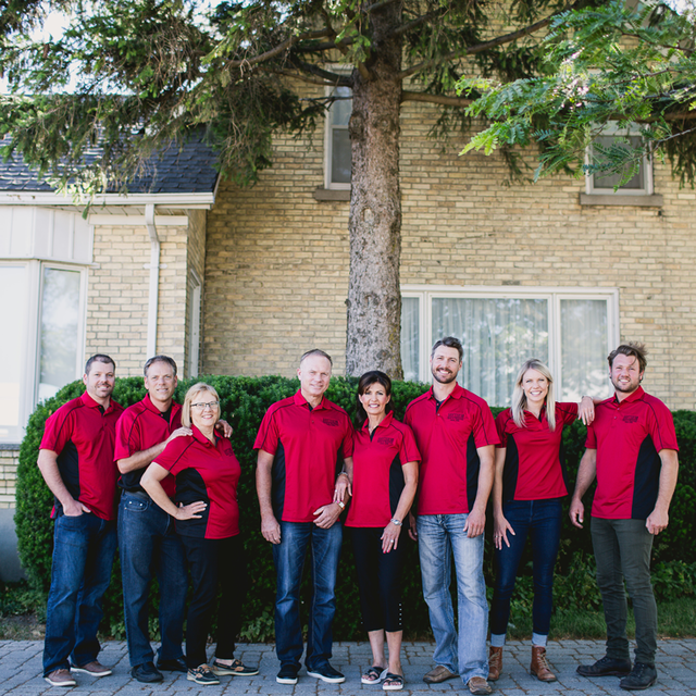 Group of eight people wearing matching red shirts standing outside a brick house.