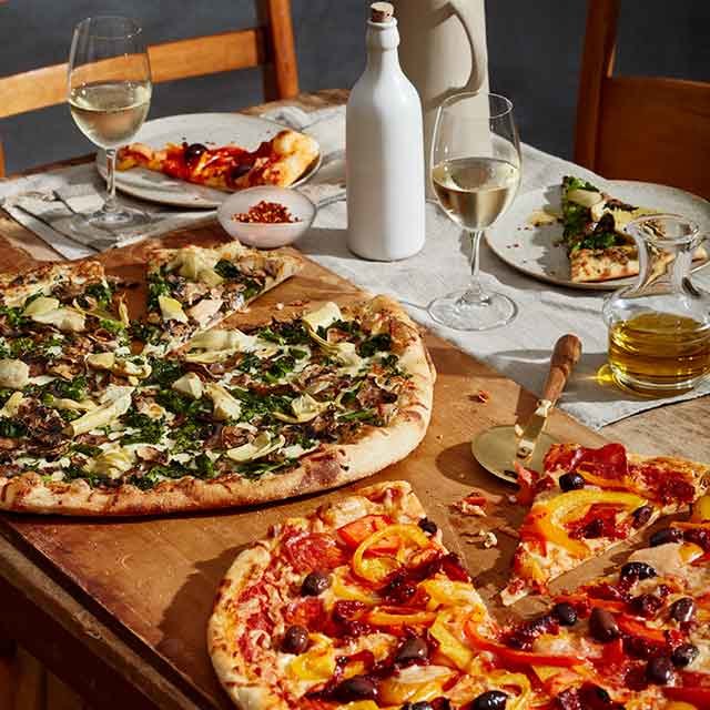 Two large pizzas on a wooden board with wine glasses, olive oil, and plates on a dining table.