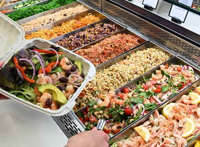 Person holding a takeout container of salad at a self-serve bar with assorted grains, beans, and vegetables.