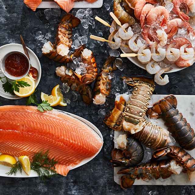 Fresh seafood display with salmon fillet, lobster tails, shrimp skewers, and cocktail sauce on ice.