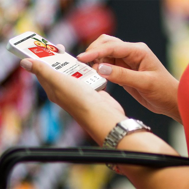 Person holding a smartphone in a grocery store, screen showing &lsquo;HELLO FRESH FOOD&rsquo; while shelves of products are blurred in the background.