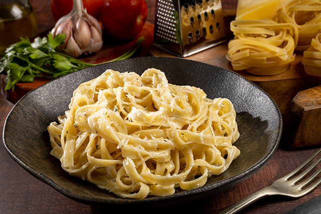 Creamy fettuccine Alfredo served in a black bowl with garlic, tomatoes and parmesan in the background.