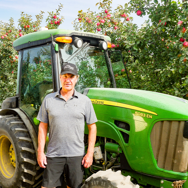 Person standing in front of a green John Deere 5083 EN tractor in an apple orchard.