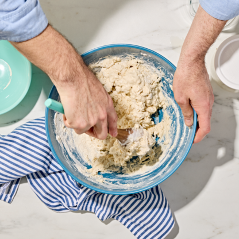 Two hands mixing a Cherry Chocolate Sourdough Loaf starter in a blue bowl on a marble surface.